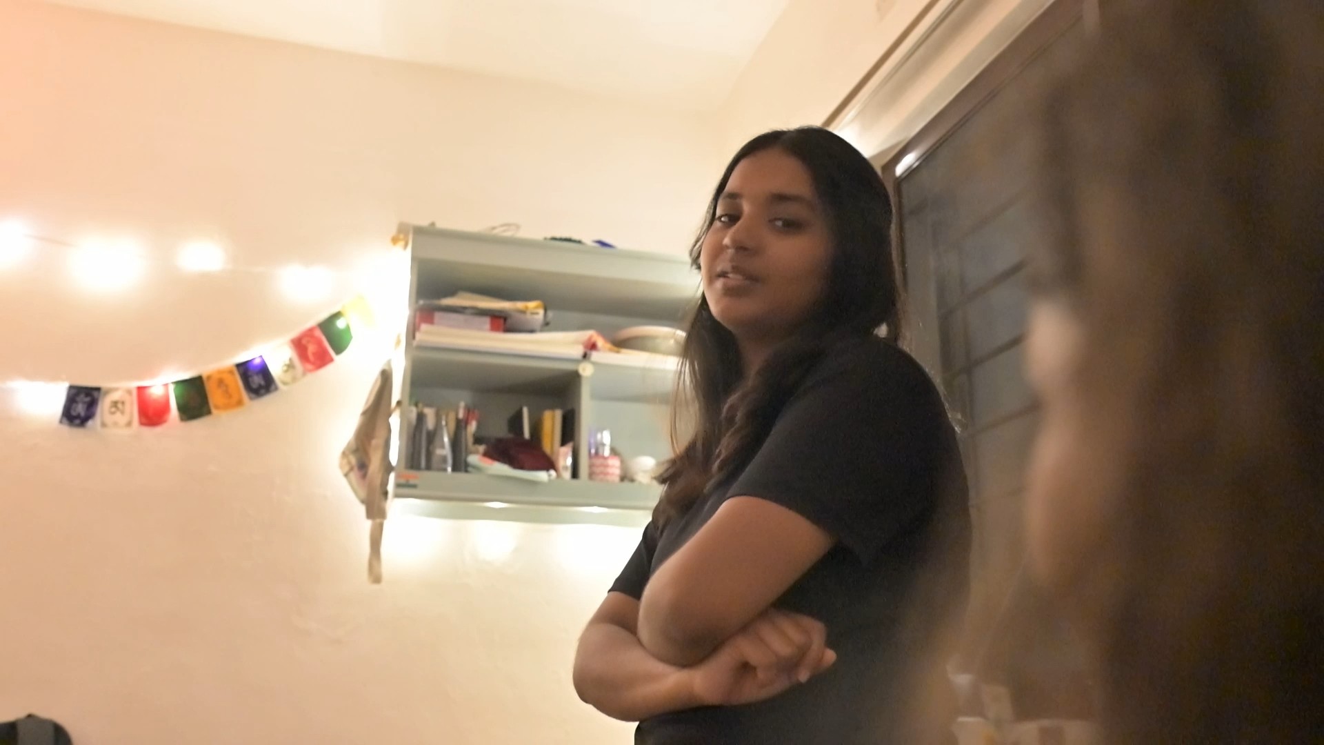 A girl in a room lit by fairy lights, arms crossed, looking back at the camera